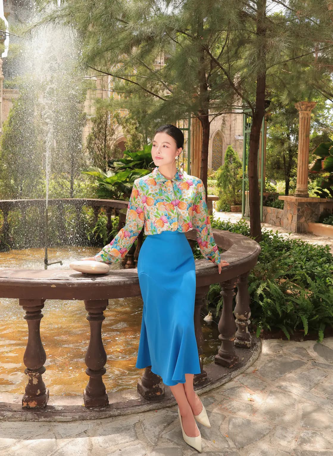 Người mẫu ngồi bên đài phun nước trong sơ mi hoa và chân váy xanh.